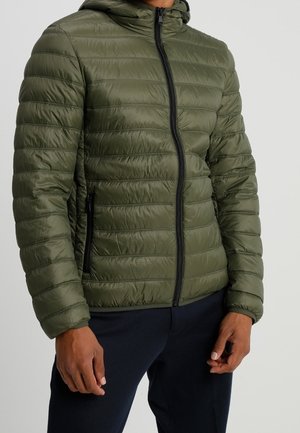 Homme portant une veste matelassée à capuche de couleur vert olive avec une fermeture éclair noire et des poches latérales, associée à un pantalon foncé.