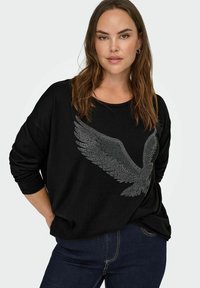 Black long-sleeve sweatshirt featuring a silver jeweled eagle design. Relaxed fit with rolled cuffs and a round neckline. Paired with dark jeans.