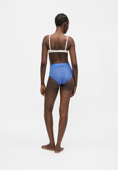 Γυναίκα με πλεξούδες στέκεται ξυπόλητη με την πλάτη, φοράει λευκό σουτιέν και μπλε εσώρουχο με ψηλή μέση πάνω σε απλό φόντο.