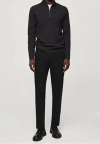 Jersey de cuello con cremallera de color gris oscuro, confeccionado en tejido de punto, combinado con pantalones negros de corte ajustado y zapatos negros, posando contra un fondo neutro.