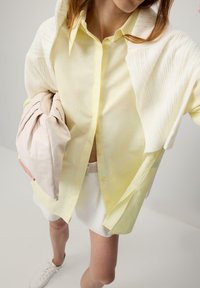 Cămașă galbenă cu nasturi, overlay alb din tricot, geantă crem cu pliuri și pantaloni scurți albi. Ținuta are tonuri deschise și o croială relaxată.