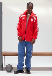 Raudona striukė užtraukiama su baltomis „Adidas“ juostelėmis, pilka marškinėliai, mėlyni džinsai su šoninėmis juostelėmis ir juodi batai. Šalia yra pilkas krepšinis.