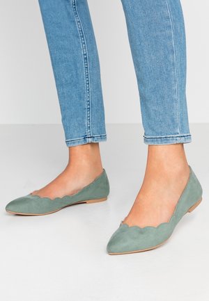 Jasnoniebieskie dżinsy w połączeniu z szałwiowymi zamszowymi balerinami w kształcie falbany na białej podłodze.