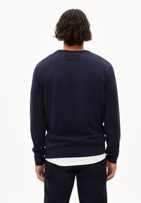 Navyblauwe gebreide trui met lange mouwen, textuurstof en ronde halslijn. Bevat een klein label aan de achterkant. Draagt over een wit shirt.