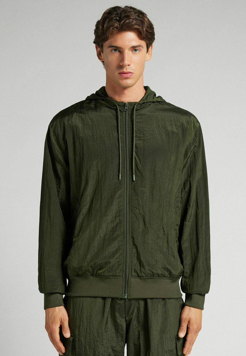 Chaqueta ligera color verde oliva con capucha y textura arrugada, con cremallera frontal y puños acanalados. Diseño minimalista, sin patrones ni acentos.