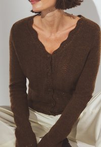 Bruine gebreide cardigan met een golvende halslijn, knoopsluiting en lange mouwen. Zachte textuur met een delicate, open breipatroon.