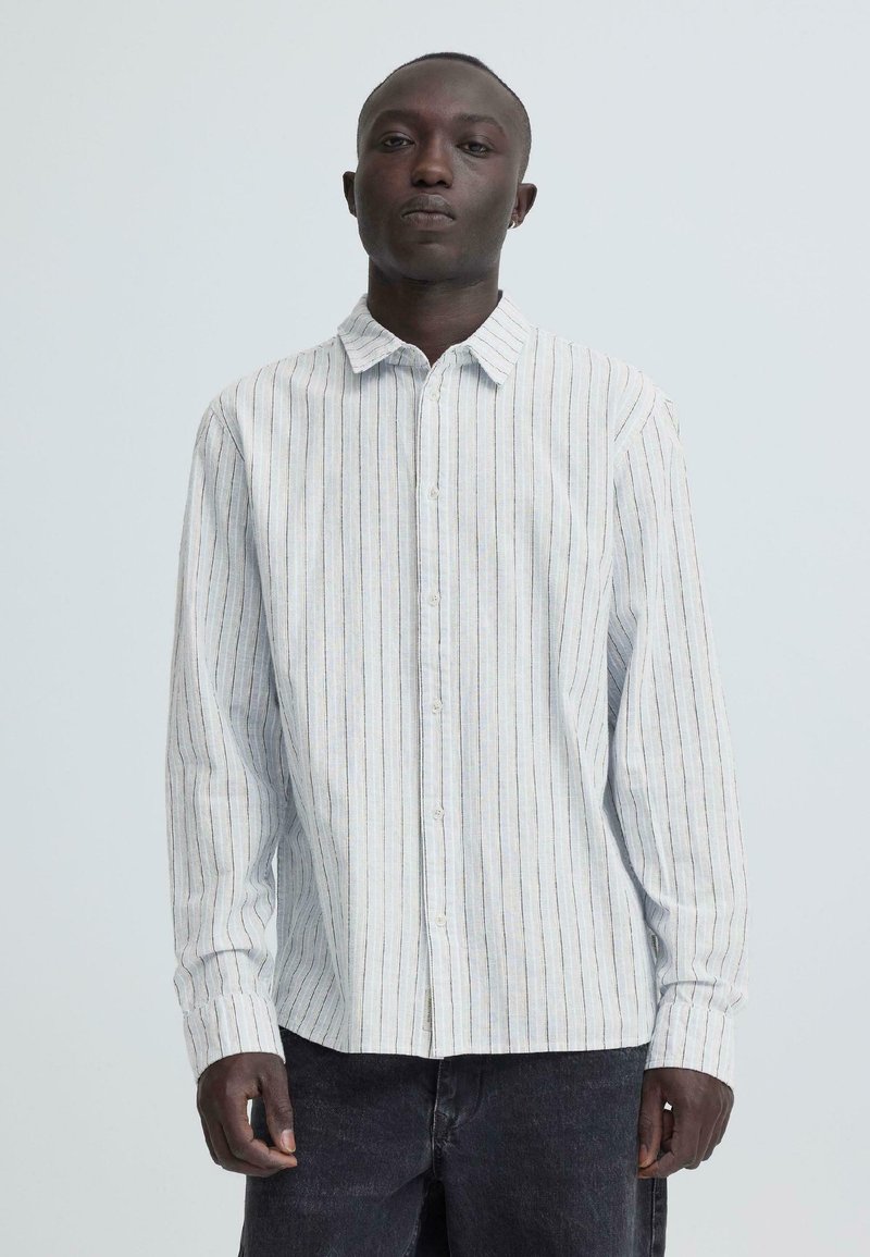 Άνδρας που φοράει λευκό πουκάμισο με μακριά μανίκια, κουμπωτό, με λεπτές κάθετες ρίγες και σκουρόχρωμο παντελόνι, μπροστά σε απλό φωτεινό φόντο.