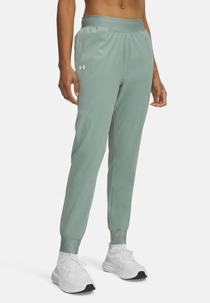 Personne portant un pantalon de jogging Under Armour vert clair et des chaussures de sport blanches, debout devant un fond clair uni.
