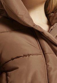 Brun, quiltet jakke med hætte og en blød, glat tekstur. Har en lynlås foran og en tætsiddende pasform.