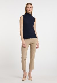 Top tricot fără mâneci de culoare bleumarin, cu guler înalt, asociat cu pantaloni bej tapered și pantofi cu toc nude. Textură netedă și design adaptat.