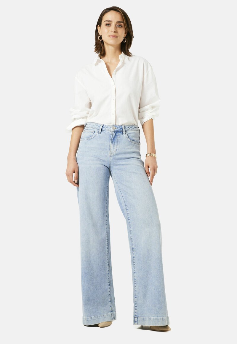 Vrouw staat in een witte blouse met opgerolde mouwen, lichtblauwe high-waisted wijde jeans en beige schoenen.