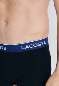 Boxer ajustados negros con una cinturilla azul marino que muestra "LACOSTE" en letras blancas. Tela suave y diseño ceñido.