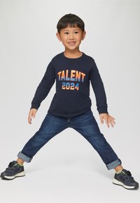 Maglietta a maniche lunghe della marina con "TALENT 2024" stampato in arancione e blu. Indossata con jeans in denim scuro arrotolati e scarpe da ginnastica nere sportive.