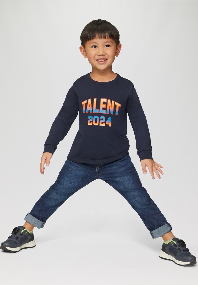 Maglietta a maniche lunghe della marina con "TALENT 2024" stampato in arancione e blu. Indossata con jeans in denim scuro arrotolati e scarpe da ginnastica nere sportive.