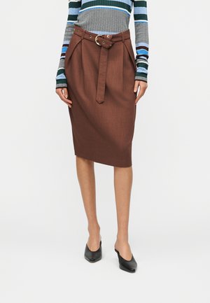 Mujer con falda marrón hasta la rodilla con cinturón, camiseta de manga larga a rayas y zapatos negros de tacón, de pie contra un fondo liso.