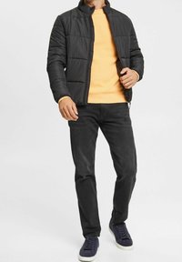 Schwarze gesteppte Jacke, orangefarbenes Langarmshirt, schwarze Jeans und marineblaue Turnschuhe. Die Jacke hat einen Stehkragen und keine sichtbaren Logos oder Muster.