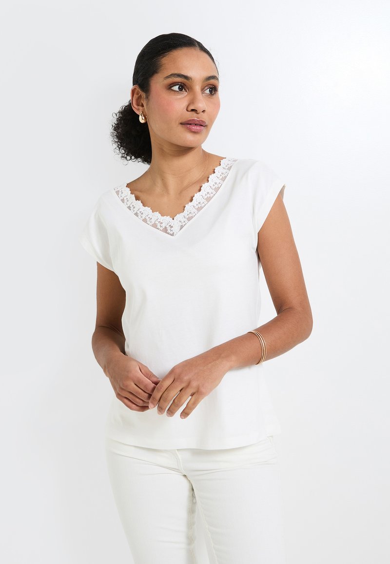 Haut blanc à manches courtes avec un décolleté en V orné d'une bordure en dentelle festonnée. Tissu doux, coupe décontractée, associé à un pantalon blanc.