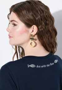 Námořnické tričko s krátkými rukávy, s bílým vyšitým textem "dive with the flow" a motivy ryb na zádech. Model má na sobě zlaté náušnice.