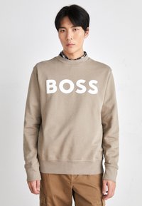 Joven vestido con un sudadera beige con "BOSS" en letras blancas, una camisa a cuadros debajo y pantalones marrones, de pie frente a un fondo liso.