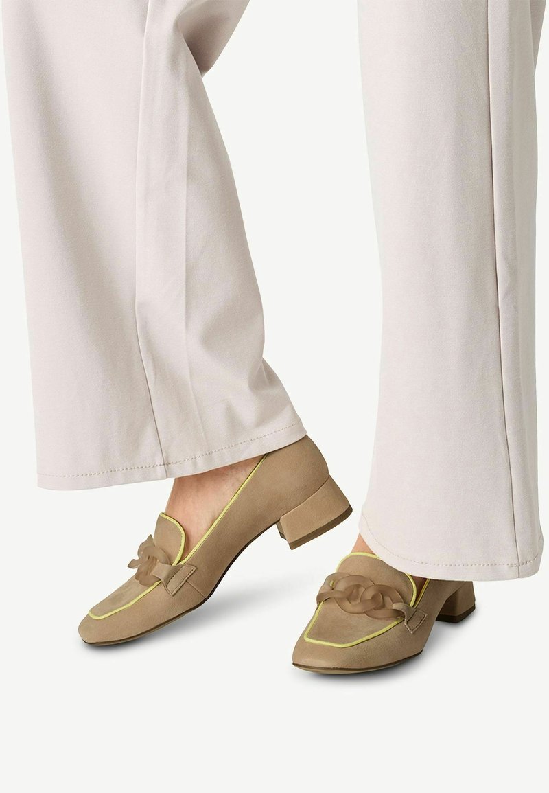 Chaussures à talons carrés en daim beige avec un liseré jaune et un nœud décoratif sur chaque orteil. Portées avec un pantalon pale à jambes larges, masquant partiellement les chaussures.