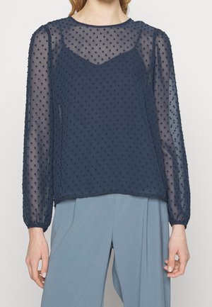 Vrouw draagt een doorschijnende marineblauwe polkadotblouse met lange mouwen over een hemdje, gecombineerd met lichtblauwe hoge taillebroeken.