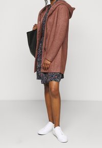 Cardigan lungo color ruggine con cappuccio, caratterizzato da una finitura testurizzata e chiusura con bottoni, indossato sopra un vestito floreale e abbinato a sneakers bianche.