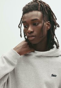 Tânăr cu dreadlocks purtând un hanorac gri deschis, privind în jos și ajustând gulerul hanoracului cu o mână.