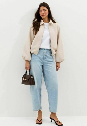 Vrouw die een beige jack met rits draagt, een wit shirt, lichtblauwe hoge taille jeans, zwarte sandalen met hak, en een kleine donkerbruine handtas vasthoudt.