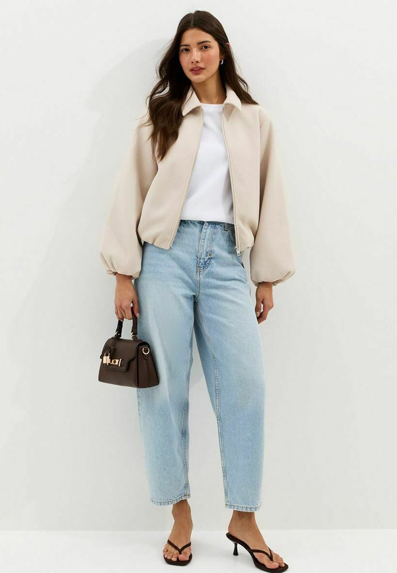 Mujer con chaqueta beige con cremallera, camiseta blanca, vaqueros de tiro alto azul claro, sandalias negras de tacón y un pequeño bolso marrón oscuro en la mano.