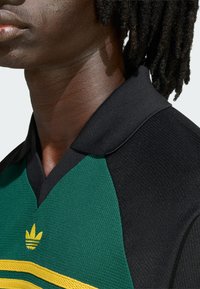 Crna sportska majica s ovratnikom u zelenoj boji s žutim prugama, izrađena od teksturiranog materijala s izvezenim logotipom na prsima.