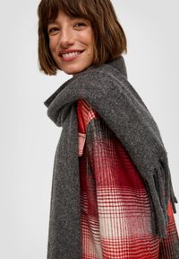 Grau gestrickter Schal mit Fransen, der über einem rot-weißen karierten Hemd hängt. Der Schal ist weich und strukturiert und sorgt für Wärme im geschichteten Outfit.