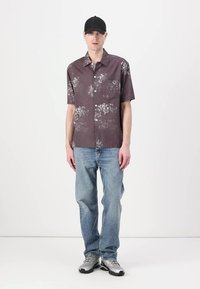 Chemise à manches courtes, à motifs floraux maroon, avec une poche poitrine, accompagnée de jeans bleu clair et de chaussures de sport grises. Casquette noire portée.