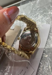 Gold-tone wristwatch with a patterned metal band and a round case. Features a glass back displaying branding and water resistance details.