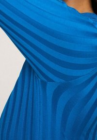 Blau gestreiftes Oberteil mit abwechselnd helleren und dunkleren Streifen. Figurbetonter Schnitt, weicher Stoff und runde Kragenlinie, die auf der Schulter sichtbar ist.