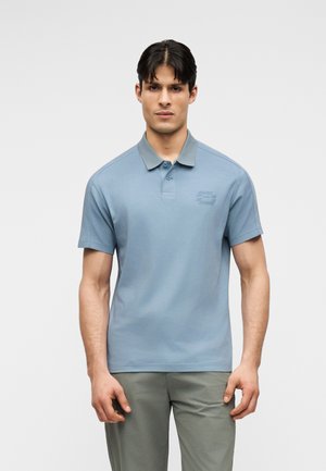Man draagt een lichtblauw poloshirt met korte mouwen, een grijze kraag en grijze broek, staand tegen een effen lichte achtergrond.