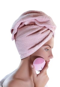 Serviette rose enroulée autour des cheveux, tenant une brosse nettoyante faciale en silicone rose avec une surface texturée, sur fond blanc.