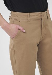 Chino en coton couleur kaki avec une coupe slim, présentant un devant plat, des passants de ceinture et des poches latérales. Texture lisse avec une taille régulière.
