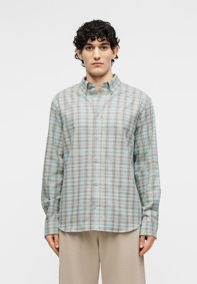 Νεαρός άνδρας που φοράει ανοιχτό μπλε και μπεζ πουκαμίσα με κουμπιά και μπεζ παντελόνι, στέκεται ανάμεσα σε απλό φόντο, κοιτάζοντας προς τα εμπρός.