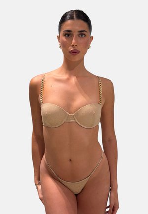 Donna con capelli scuri in bikini dorato con spalline a catena, che guarda avanti su uno sfondo chiaro e uniforme.