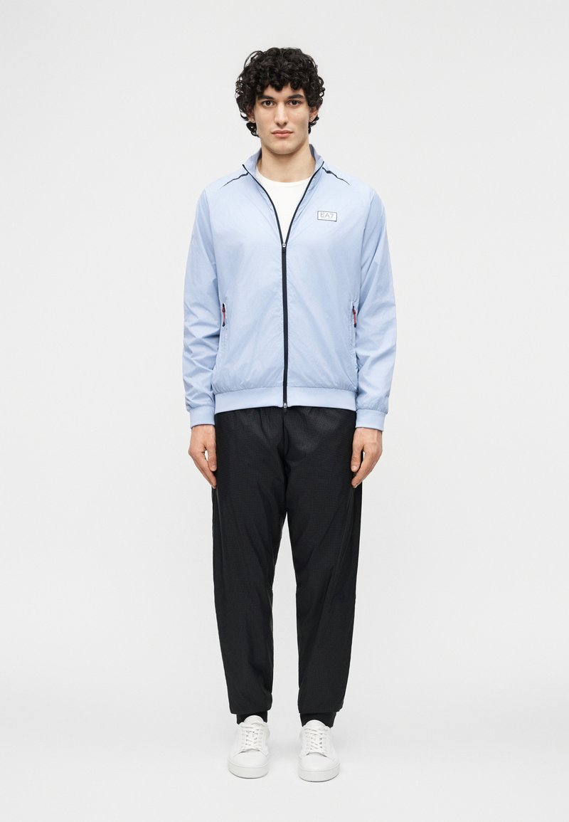 Νεαρός με σγουρά μαλλιά που φορεί ανοιχτό γαλάζιο μπουφάν με φερμουάρ, μαύρο παντελόνι και λευκά αθλητικά παπούτσια, στέκεται μπροστά σε ένα απλό λευκό φόντο.