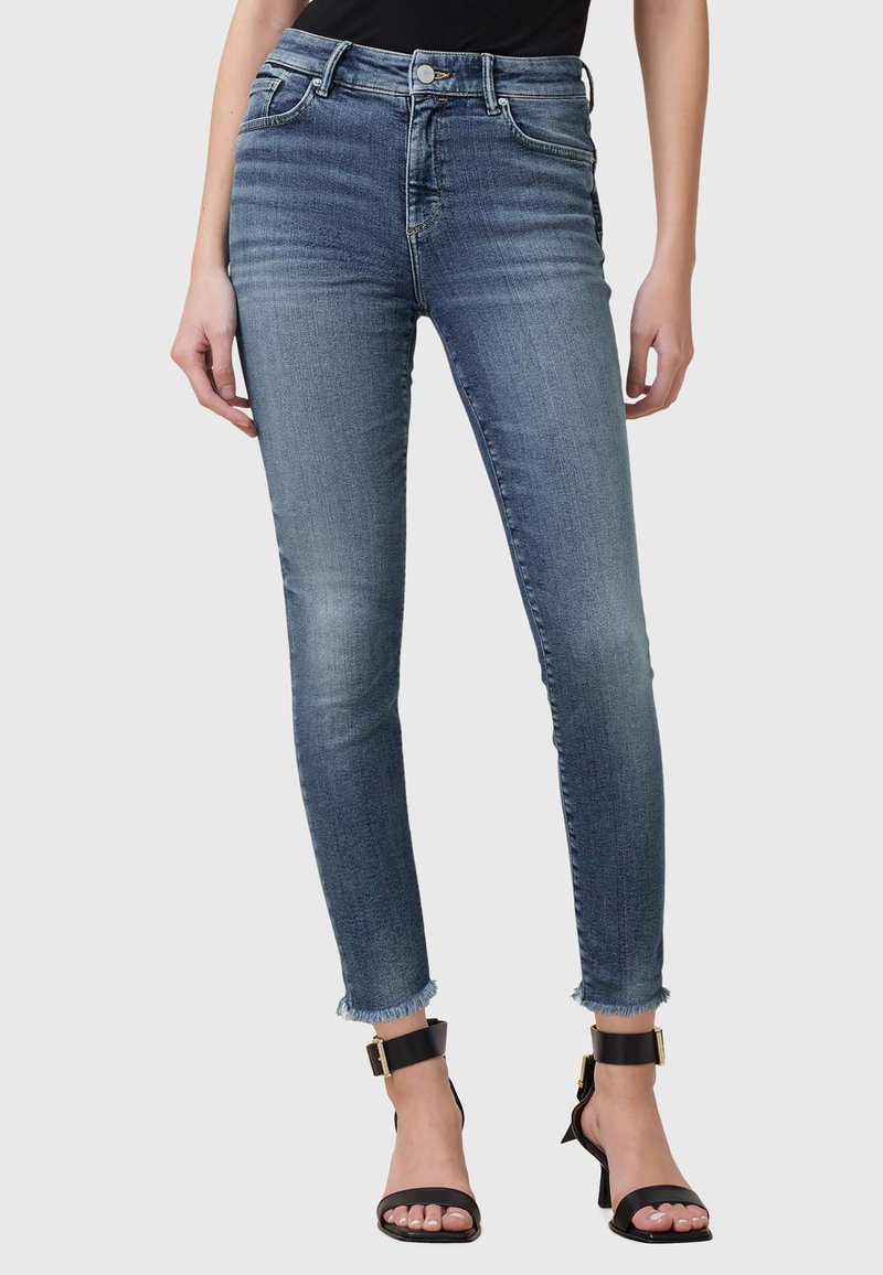 Chude dżinsy w średnim niebieskim kolorze, z wysokim stanem, pięcioma kieszeniami i surowym wykończeniem. Zestawione z czarnymi sandałami na kostkę.