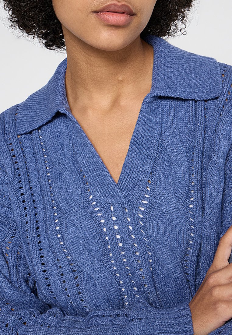 Donna che indossa un maglione azzurro lavorato a maglia con motivi a treccia e un ampio colletto, braccia incrociate, labbra chiuse.