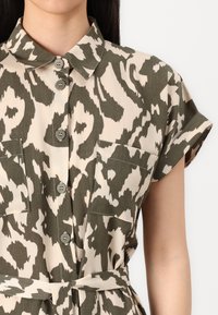 Abito camicia beige con motivo astratto verde, maniche corte, colletto e due tasche sul petto. Include una cintura di tessuto in vita.