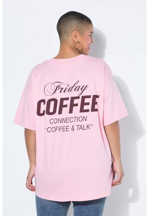Person med kort hår iført en lyserød, oversized t-shirt med teksten "Friday COFFEE CONNECTION 'COFFEE & TALK'" på ryggen, stående mod en hvid væg.
