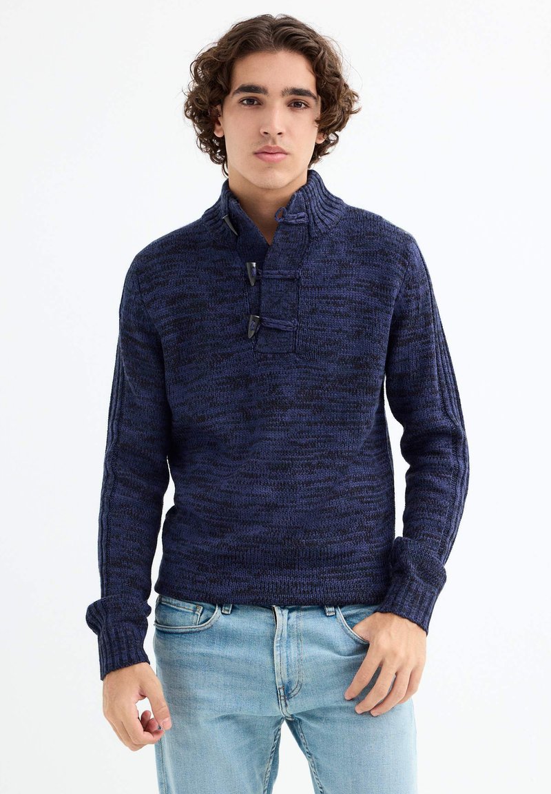 Suéter de punto azul marino con un cuello alto, que presenta tres botones y puños acanalados. Combinado con jeans de mezclilla azul claro.