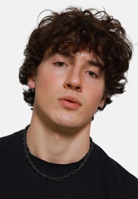 Collana nera a catena con anelli metallici intrecciati. Indossata sopra una t-shirt nera, con una texture della pelle liscia e capelli scuri arruffati.