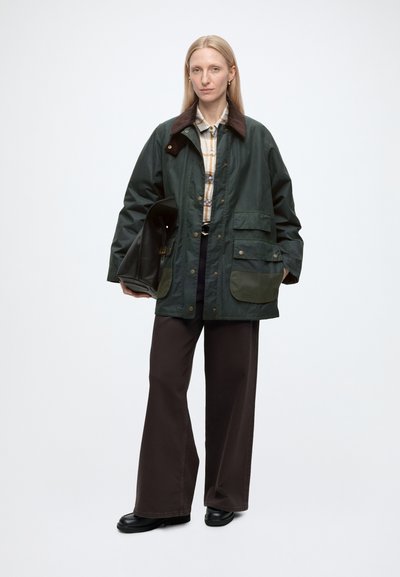 Femme aux longs cheveux blonds porte une veste vert foncé sur une chemise à carreaux et un pantalon large foncé, tenant un grand sac noir sur fond blanc.