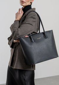 Sac fourre-tout en cuir noir avec finition texturée, doté de deux poignées robustes et d'un design minimaliste. Accent en logo doré à l'avant.