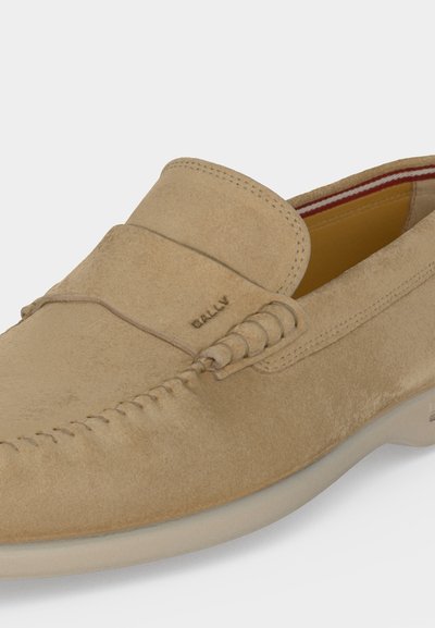 Μπεζ μοκασίνι slip-on από σουέντ με ραφές μοκασίνου, λαστιχένια σόλα και κόκκινη ριγωτή εσωτερική φόδρα.