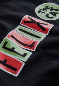 Črn material z barvitim, blokovnim besedilom. "FELIX" v rdeči in zeleni barvi, obrobljen v beli. Krožni poudarek nosi napis "Mačka" v zeleni barvi z black tekstom.